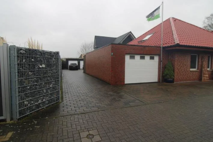 Zufahrt mit Pflasterung, Gabionenzaun und Garage, dahinter Wohnhaus mit roten Dachziegeln.