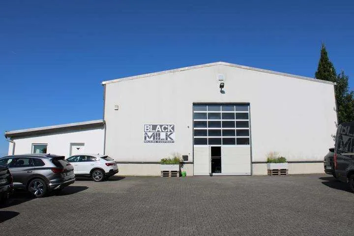 Frontansicht einer weißen Gewerbehalle mit Torelement und Parkplatz davor.