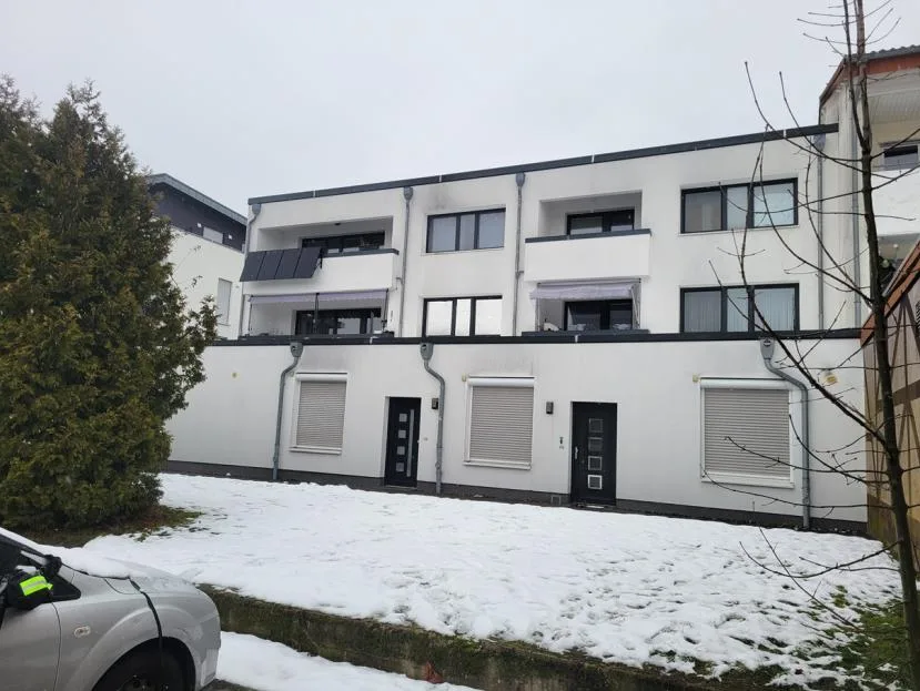 Rückansicht eines weiß verputzten Mehrfamilienhauses mit Balkonen, davor verschneiter Garten und ein parkendes Auto.