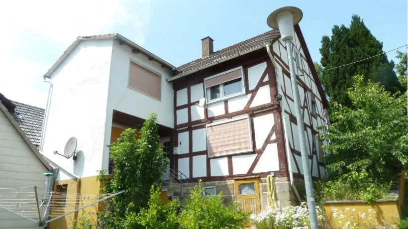 Fachwerkhaus mit weißer Putzfassade, frontale Außenansicht mit Garten und Vordach, Gebäude gut sichtbar.