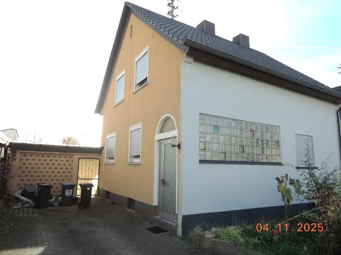 Wohnhaus mit gelber und weißer Fassade, Satteldach und Glasbausteinfenster, Eingang und Zufahrt mit Mülltonnen.