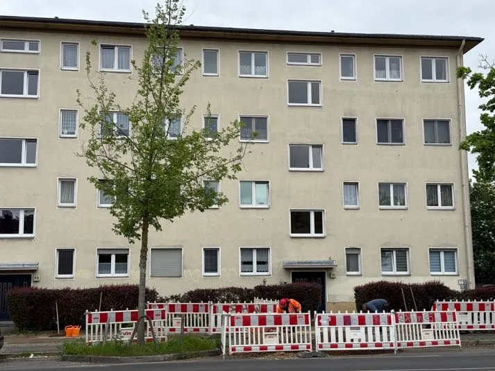Frontalansicht eines viergeschossigen Wohngebäudes mit gleichmäßiger Fensterreihe, teilweise von Baum und Absperrung verdeckt.