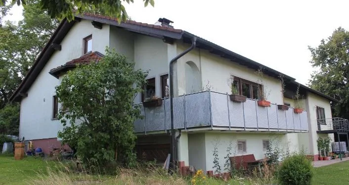 Wohngebäude mit Balkon und Gartenbereich, teilweise von einem Baum verdeckt, schräge Seitenansicht.