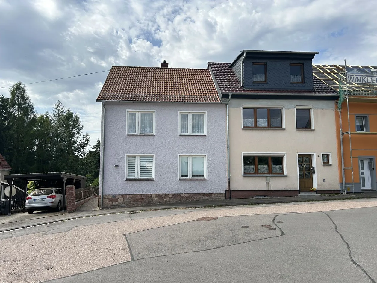 Straßenansicht eines Doppelhauses mit verputzter Fassade, Dachgaube und Carport links im Bild.
