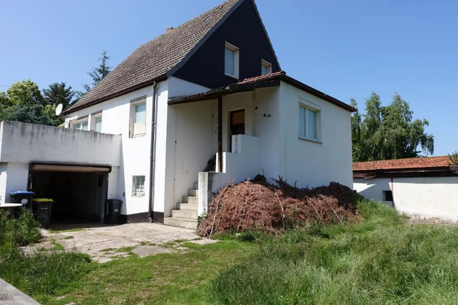Einfamilienhaus mit dunklem Dachgiebel, weißer Fassade und Garageneinfahrt, von vorne-seitlich aufgenommen.