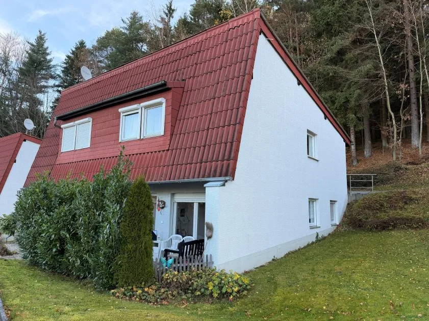 Frontansicht eines Einfamilienhauses mit roter Dachflaeche und weisser Fassade, umgeben von Baumbestand.
