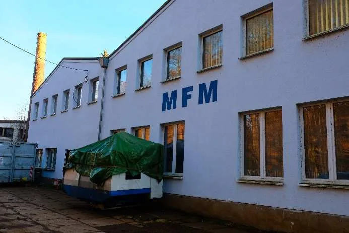 Helles zweigeschossiges Industriegebäude mit Fensterreihen, davor abgedecktes Fahrzeug auf befestigtem Hof.