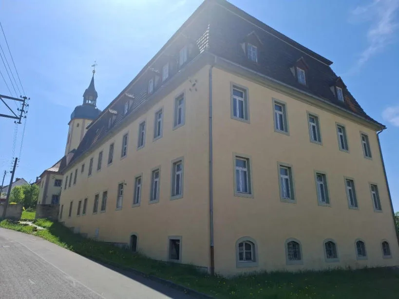 Außenansicht eines großen gelb verputzten Gebäudes mit Satteldach und Kirchturm im Hintergrund