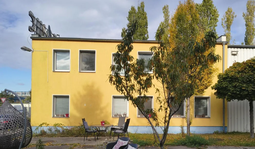 Frontalansicht des gelb gestrichenen zweigeschossigen Bürogebäudes mit Bäumen und Sitzgruppe davor