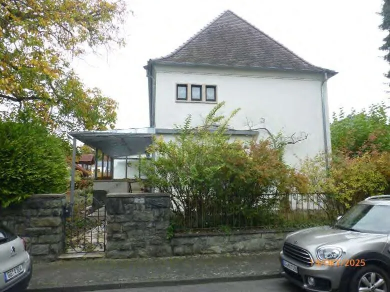 Frontansicht eines weiß verputzten Wohnhauses mit Walmdach, Vorgarten, Steinmauer und Carport.
