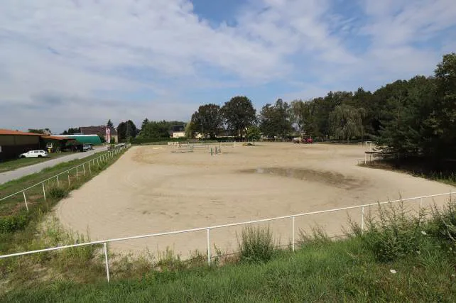 Reitplatz mit Sandboden und weißer Umzäunung, im Hintergrund Bäume und einzelne Gebäude.