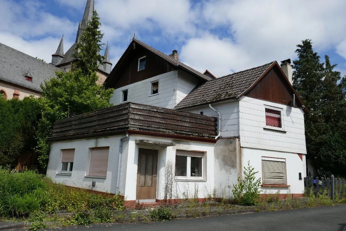 Straßenansicht des Wohnhauses mit Eingangstür, Fenstern und Holzverkleidung, im Hintergrund eine Kirche.