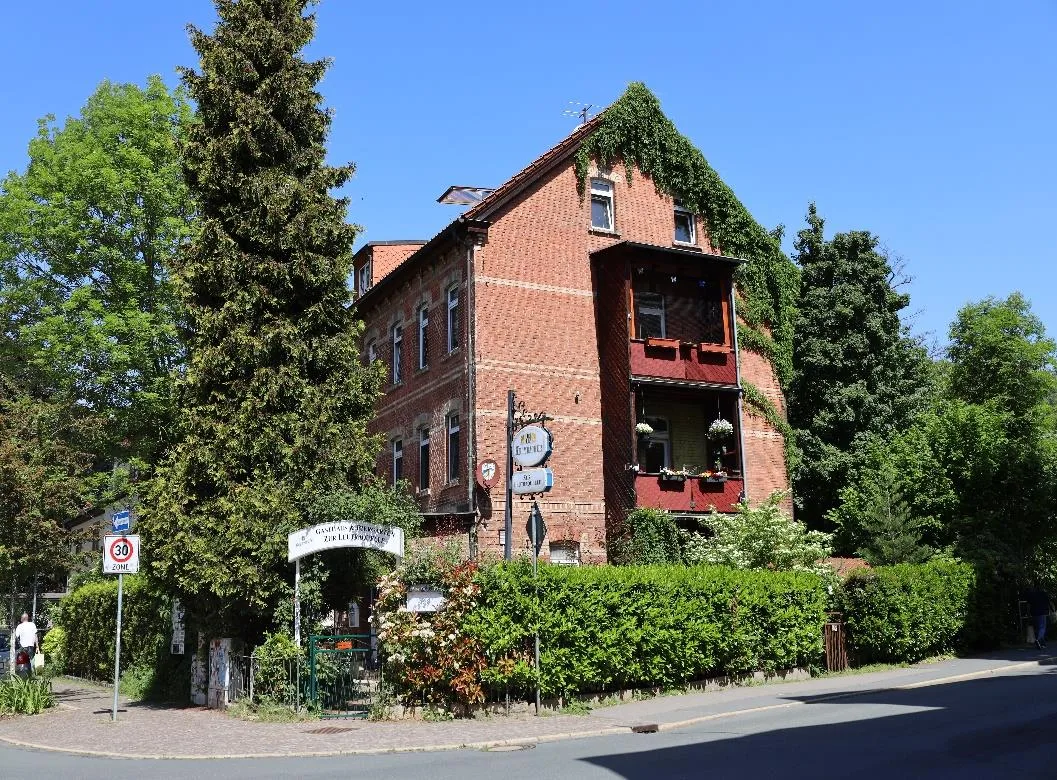 Dreigeschossiges Backsteingebäude in Eckansicht mit Balkonen, Efeubewuchs und Vorgartenhecke an der Straße, bei Sonnenschein.