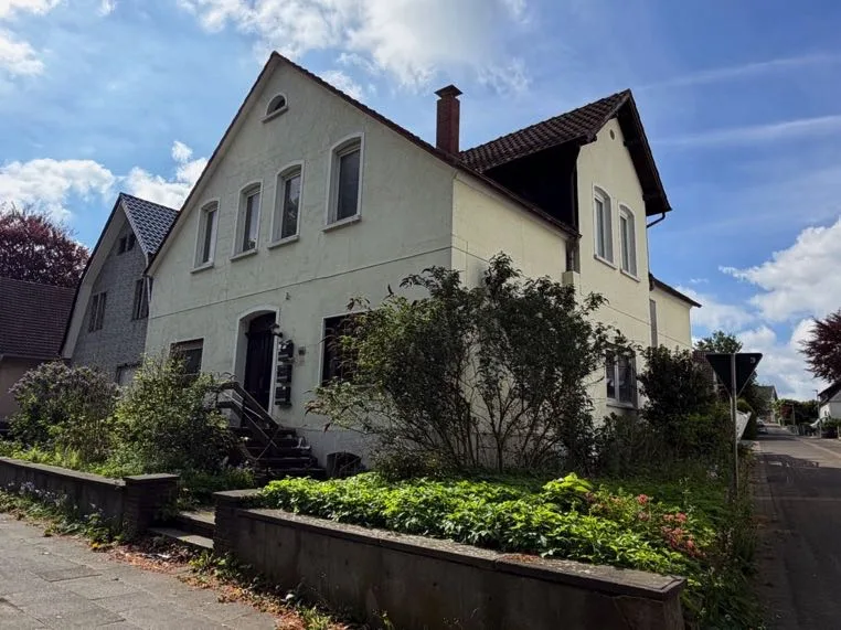 Schräge Straßenansicht eines zweigeschossigen Wohnhauses mit Satteldach und hellem Putz bei Sonnenschein.