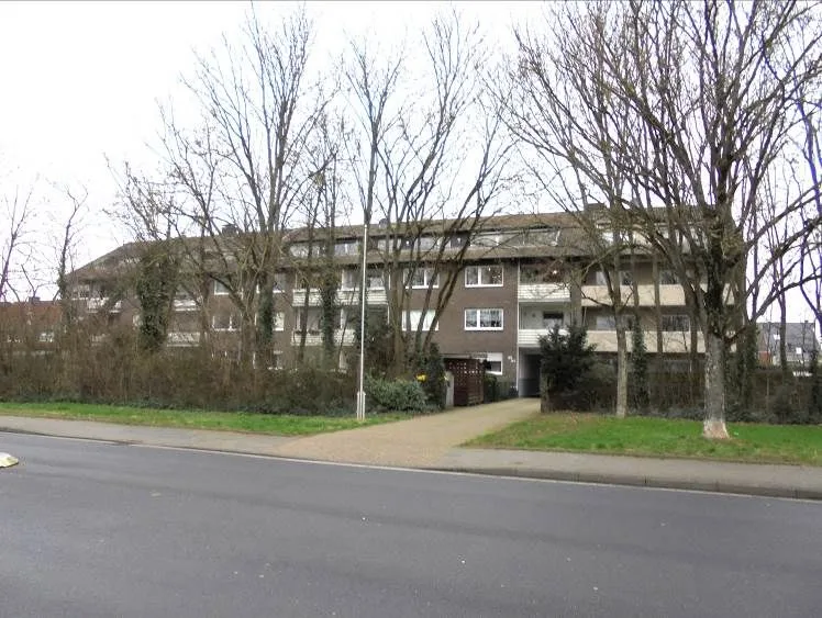 Mehrgeschossiges Wohngebäude mit Balkonen von der Straßenseite, kahle Bäume davor, Zufahrtsweg und Rasenfläche im Vordergrund.