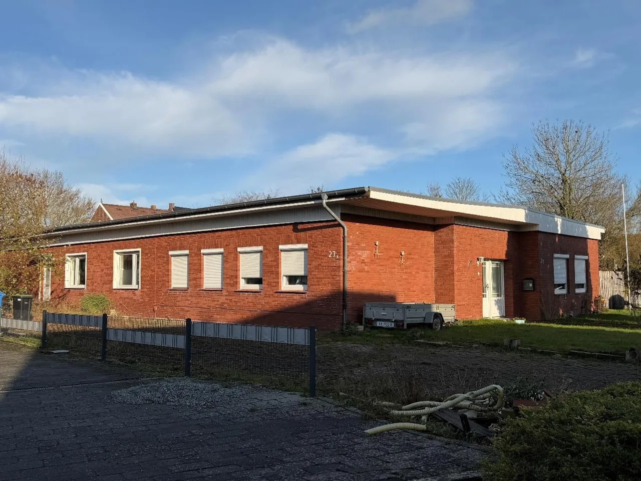 Außenansicht des eingeschossigen Klinkergebäudes mit Rollläden, Eingangstür und Vorgarten bei Sonnenlicht
