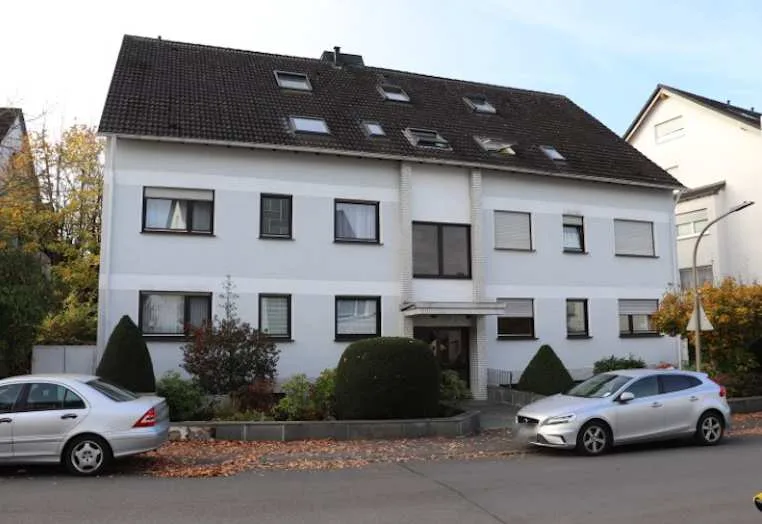 Frontansicht eines weiß verputzten Mehrfamilienhauses mit Satteldach und Dachflächenfenstern, Hauseingang mittig, Straßenansicht.