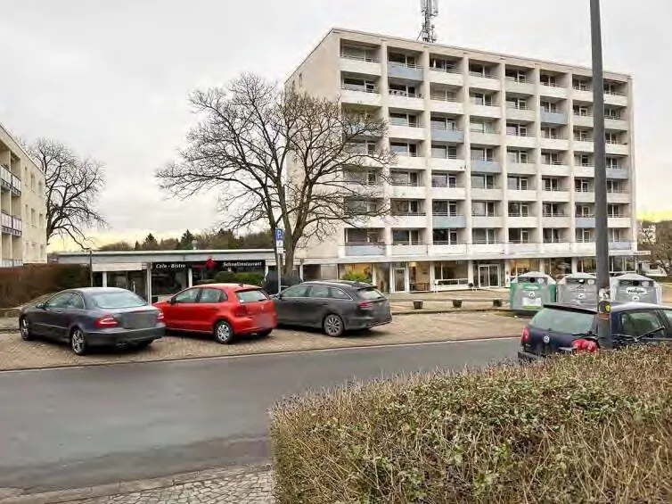 Mehrgeschossiges Wohngebäude von der Straßenseite aus, im Vordergrund parkende Autos und ein Café.