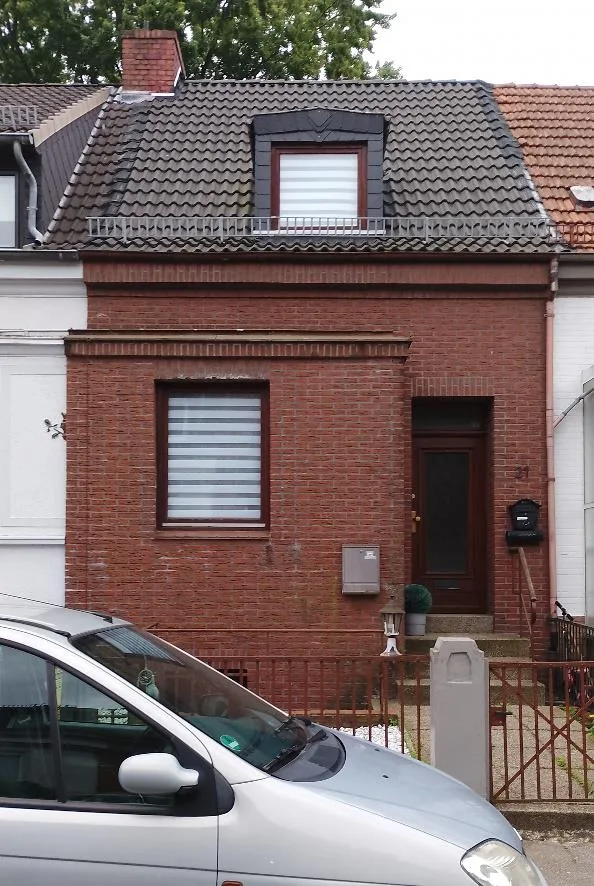 Straßenansicht: klinkerverkleidete Doppelhaushälfte mit Dachgaube, Hauseingang und Vorgarten. Ein parkendes Fahrzeug verdeckt den unteren linken Bildrand.