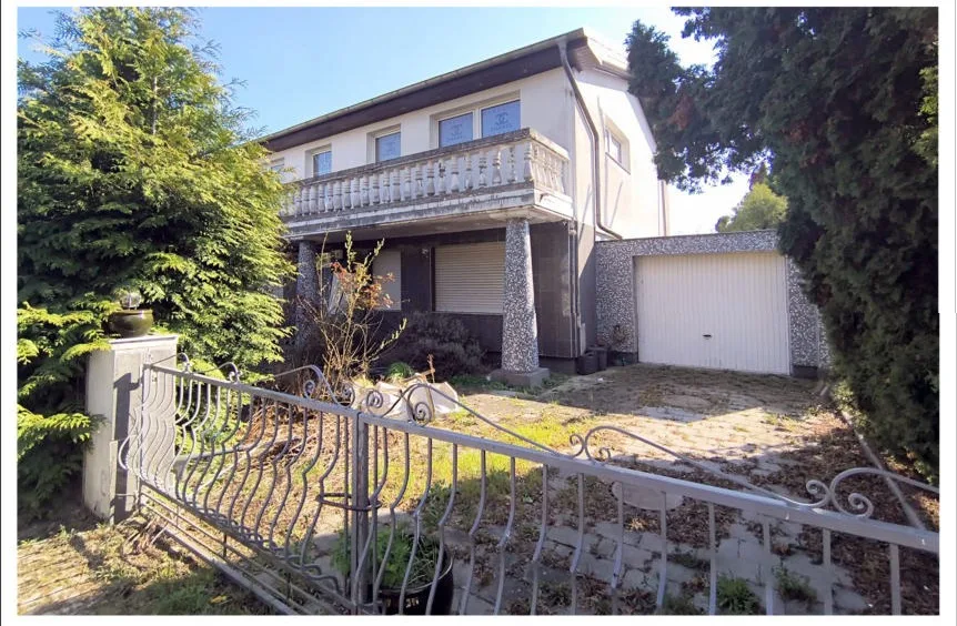 Frontansicht eines freistehenden Einfamilienhauses mit Balkon und Garage, Vorgarten mit schmiedeeisernem Zaun