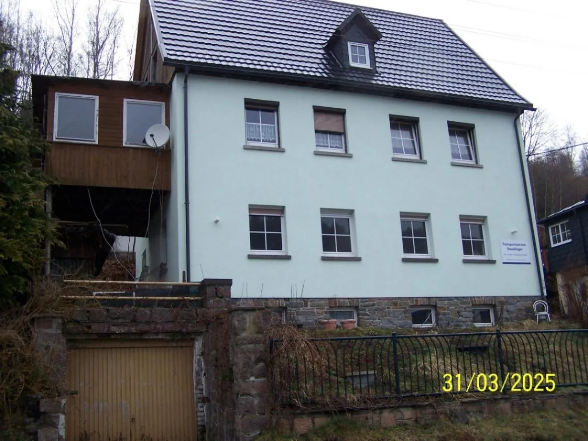 Vorderansicht eines dreigeschossigen Wohnhauses mit heller Fassade, Satteldach und Natursteinsockel; Garage im Unterbau sichtbar.