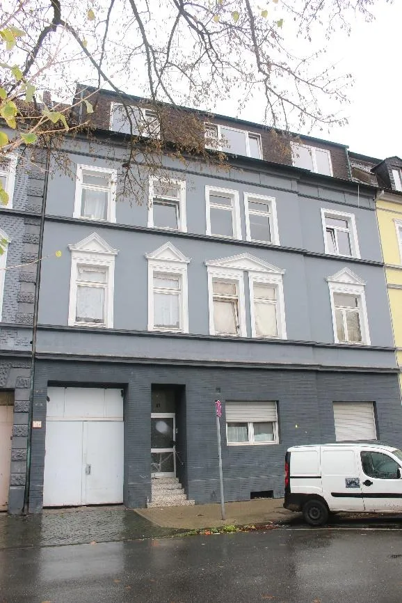 Straßenseitige Außenansicht eines mehrgeschossigen Wohnhauses mit grauer Fassade, Garagentor und Hauseingang.