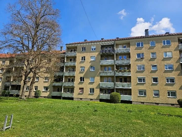 Gesamtansicht eines Wohnblocks mit Balkonen von vorne, davor Rasenfläche und Baumbestand.