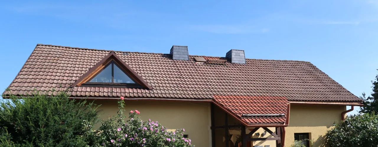 Frontansicht eines Wohnhauses mit Ziegeldach, Dachgaube und Vordach bei blauem Himmel, von Blüten gerahmt.
