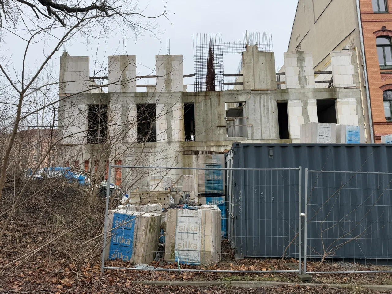 Rohbau mit Betonstützen und Ausfachungen, im Vordergrund Bauzaun, Container und Dämmstoffpaletten.