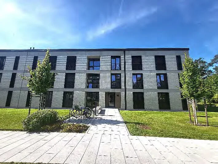 Frontale Außenansicht eines dreigeschossigen Wohngebäudes mit Eingang, Fahrradständer und Vorplatz bei Sonnenschein.