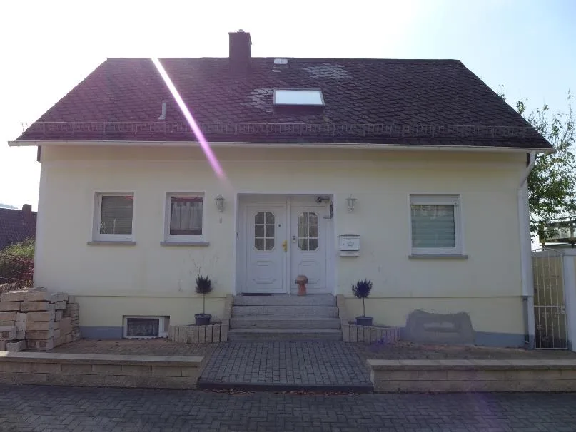 Frontansicht eines Wohnhauses mit hellgelber Fassade, mittigem Hauseingang und Treppe, Dachfenster sichtbar.
