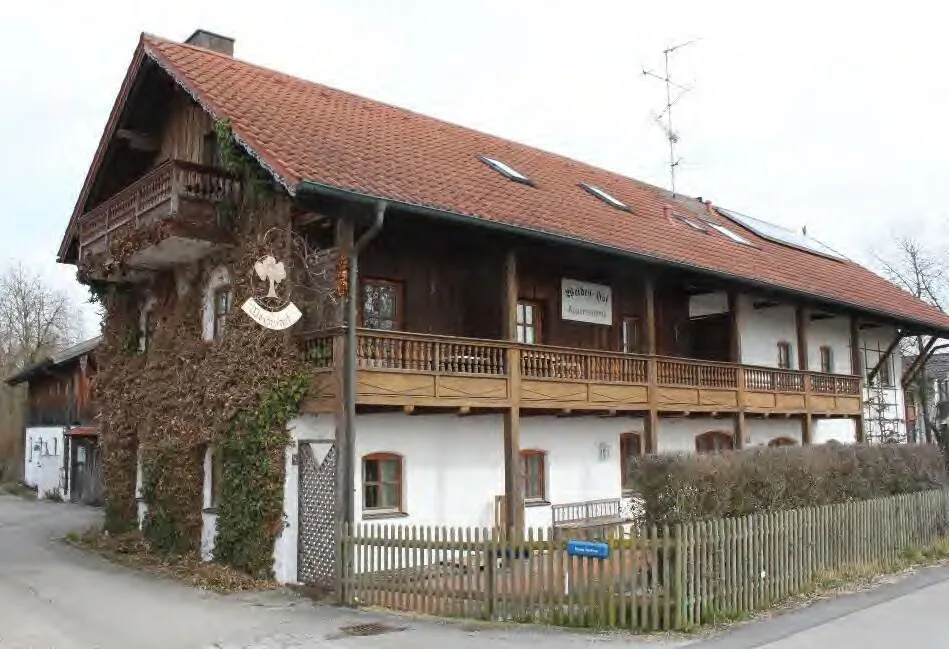 Zweigeschossiges Gebäude mit Holzbalkon und Ziegeldach, Außenansicht von vorne; Zaun und Hecke im Vordergrund.