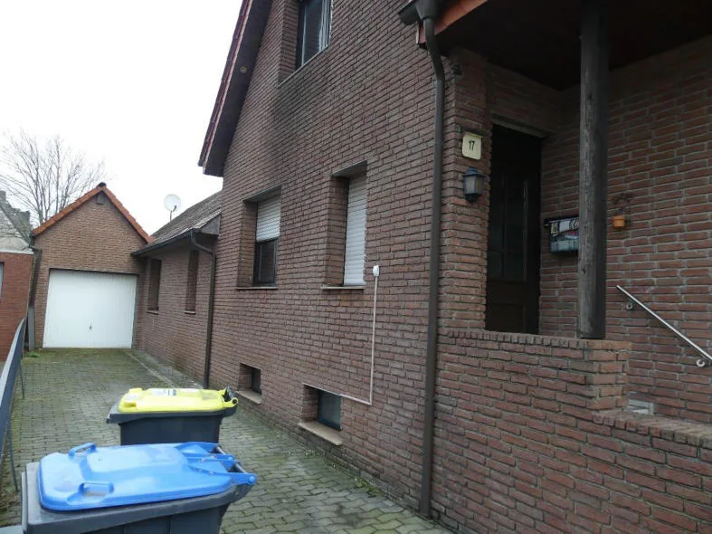 Klinkerfassade eines Wohnhauses mit Hauseingang, Garage links im Hof und Mülltonnen im Vordergrund.