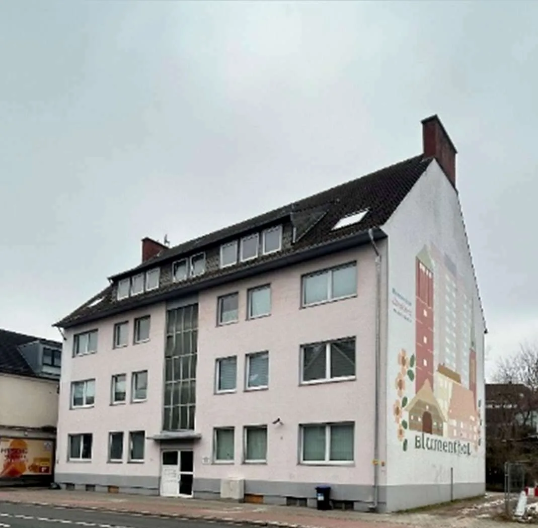 Mehrgeschossiges Wohngebäude mit heller Putzfassade, Schrägansicht von der Straßenseite, Wandbemalung an der Giebelseite.