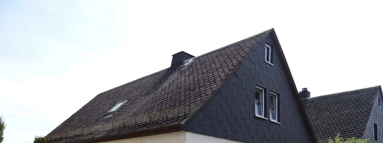 Wohnhaus mit Giebel, dunkler Schieferfassade, Dachflächen mit Dachfenstern und Schornstein vor hellem Himmel.