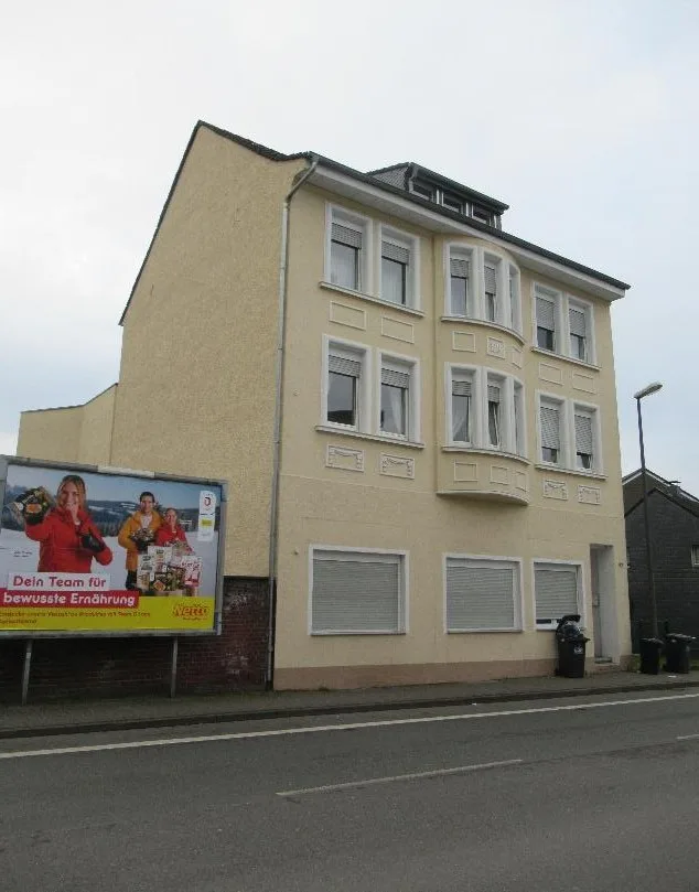 Dreigeschossiges Wohnhaus mit heller Fassade, frontale Außenansicht von der Straße aus; davor ein Werbeplakat.