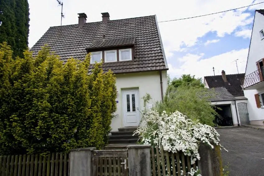 Frontansicht eines Wohnhauses mit Satteldach, Eingangstür und Treppe, teilweise von Hecke und Zaun verdeckt.