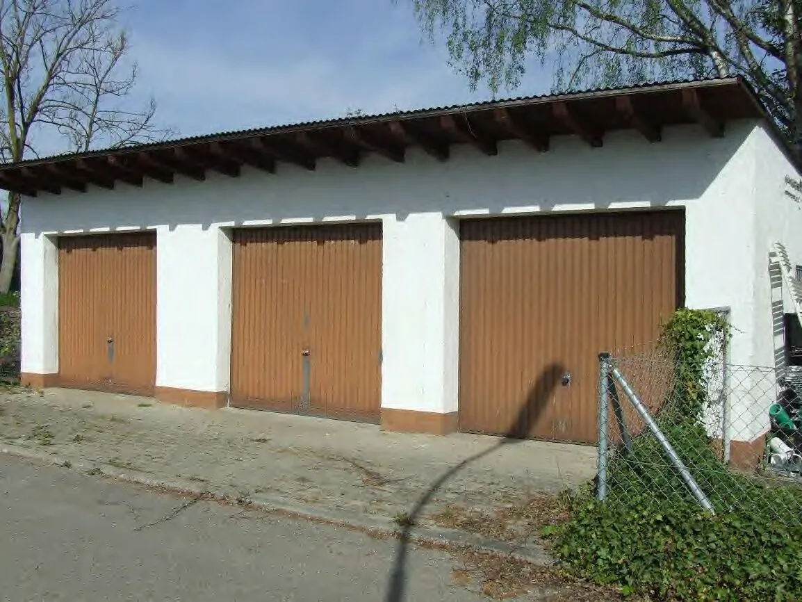 Frontansicht eines weiß verputzten Garagengebäudes mit drei braunen Schwingtoren, gut ausgeleuchtet.