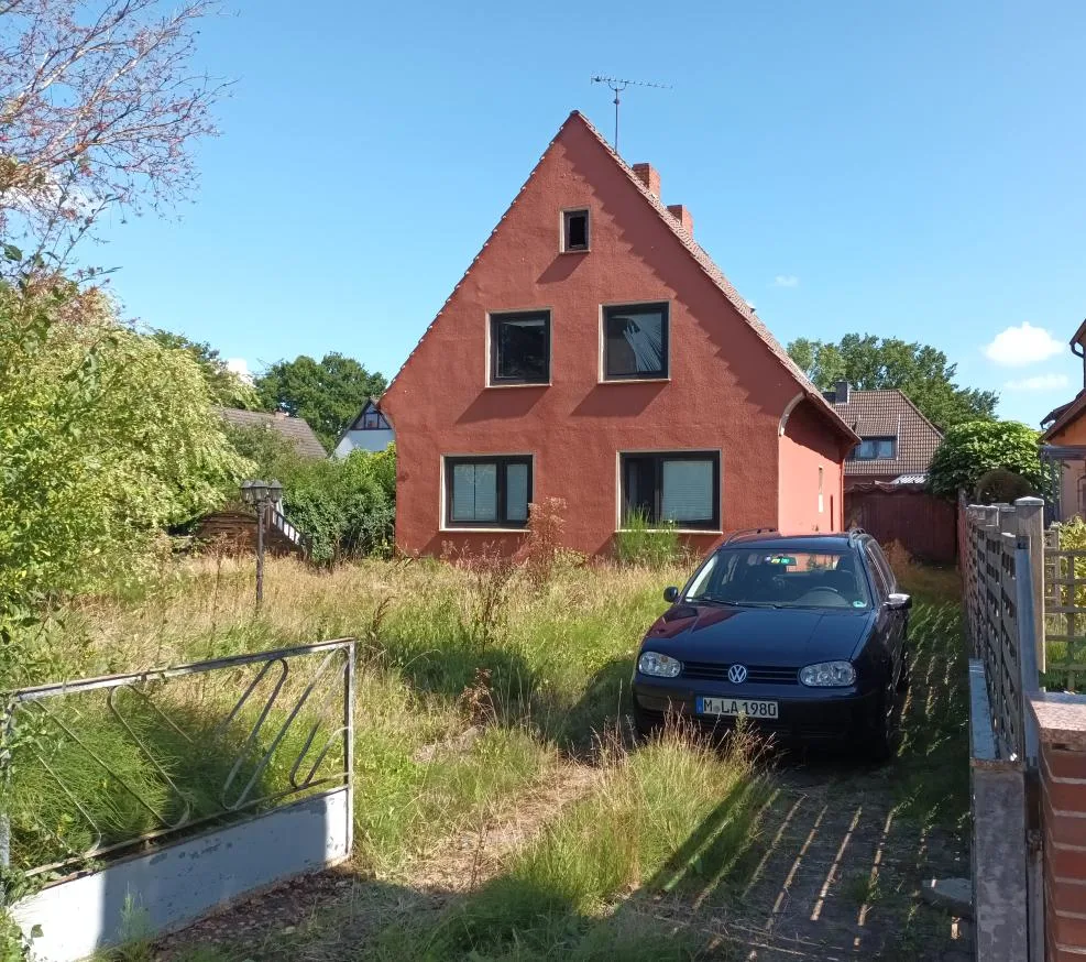 Frontansicht eines rot verputzten Einfamilienhauses mit Giebeldach, davor ungepflegter Vorgarten und Pkw.