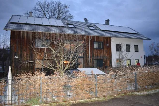 Außenansicht Wohnhaus mit Holzfassade und Photovoltaik-Dach, teilweise verdeckt durch Baum und Zaun