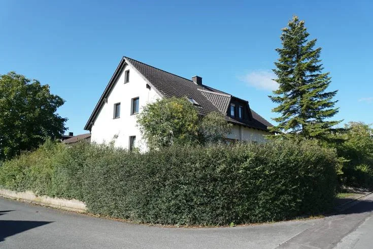 Einfamilienhaus mit weisser Fassade und dunklem Satteldach an der Strassenecke, teilweise von einer Hecke verdeckt.
