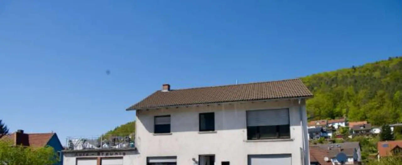 Zweigeschossiges Wohnhaus mit Ziegeldach und Rolllädenfenstern, Frontansicht vor Hanglage bei blauem Himmel.