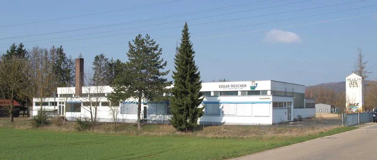 Außenansicht eines flachen weißen Betriebsgebäudes mit blauen Fensterbändern, davor Nadelbäume und Wiese, bei klarem Himmel.