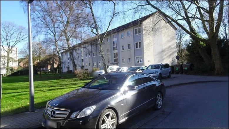 Mehrfamilienhaus mit Rasen, im Vordergrund parkende Fahrzeuge teilweise verdecken die Fassade.
