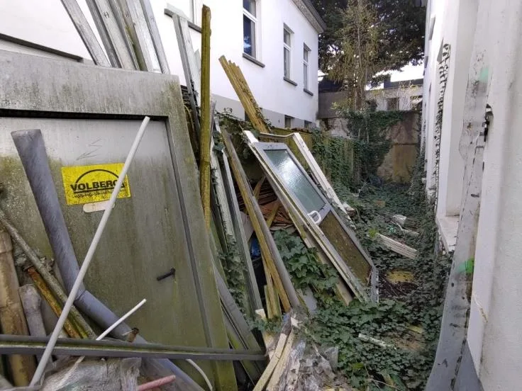 Schmaler Hof bzw. Zugang mit Bauschutt, Fensterrahmen und Bewuchs; Gebäude nur am Rand sichtbar.