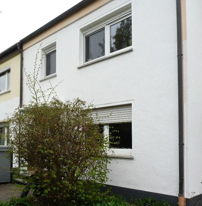 Außenansicht eines Wohnhauses mit Putzfassade und Fenstern, Gebäudeecke teilweise von einem Strauch verdeckt.