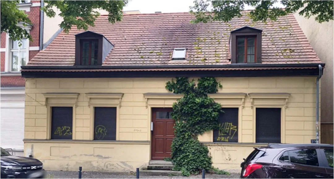 Straßenseitige Frontansicht eines zweigeschossigen Wohnhauses mit Dachgauben, Eingangstür und verschlossenen Fenstern.